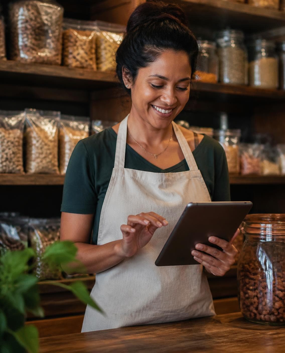 Dona de loja de produtos naturais, de avental, sorrindo enquanto usa o tablet no balcão, com prateleiras de grãos e castanhas ao fundo